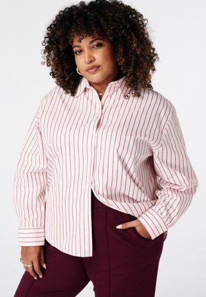 Femme aux cheveux bouclés portant une chemise à rayures rose clair boutonnée et un pantalon violet foncé, posant avec une main dans la poche sur un fond blanc.