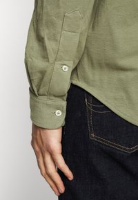 Levá ruka a ruka osoby, která má na sobě olivově zelenou košili s dlouhými rukávy a knoflíky a tmavě modré džíny se viditelným prošíváním na zadních kapsách.