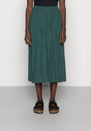 Kvinde iført en grøn plisseret midiled med sort top og sorte sandaler, stående mod en ensfarvet lys væg.