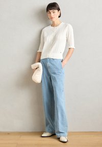 Cardigan bianco a costine con maniche corte, abbinato a jeans a palazzo a vita alta di colore blu chiaro. La modella tiene in mano una pochette color crema.
