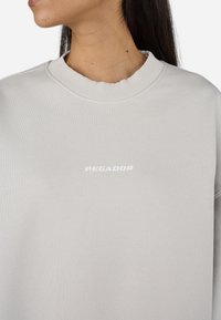 Light gray sweatshirt with a ribbed crew neckline. Features the word "PEGADOR" printed in white on the front. Soft, textured fabric.