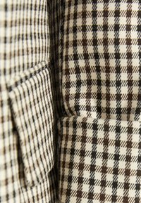 Tessuto beige con motivo a quadri marroni e neri, caratterizzato da una consistenza morbida e un design strutturato. Cuciture visibili e dettaglio di tasca.