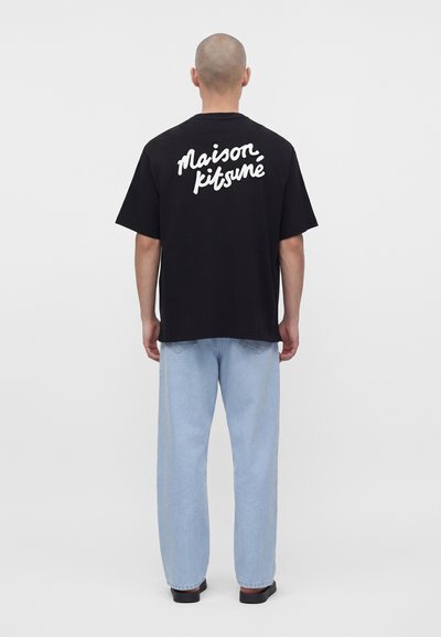 Άντρας που φοράει μαύρο T-shirt με το κείμενο «Maison Kitsuné» στην πλάτη, ανοιχτό μπλε τζιν και μαύρα παπούτσια, στέκεται γυρισμένος με την πλάτη σε λευκό φόντο.