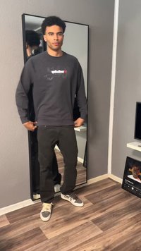 Fiatal férfi, sötét pulóvert és nadrágot visel, áll egy egész alakos tükör előtt egy fa padlós, szürke falú szobában.