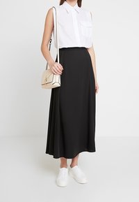 Donna che indossa una camicia bianca senza maniche con bottoni, una gonna midi nera, sneakers bianche e una borsa a tracolla beige, in piedi contro uno sfondo bianco.