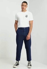 Homem em pé com as mãos nos bolsos, vestindo uma t-shirt gráfica branca, calças cropped azul-marinho e sapatilhas pretas de cano alto.