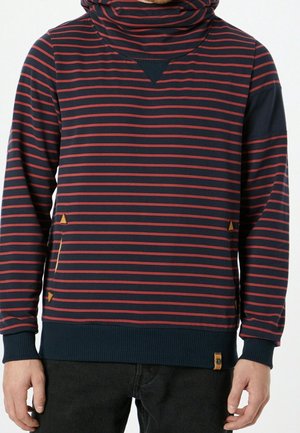 Hombre con sudadera con capucha de rayas azul marino y rojo, con puños y dobladillo acanalados, y detalles triangulares en parches de cuero marrón en los bolsillos frontales.