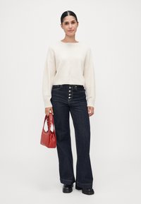 Lys beige genser, høyt liv mørkeblå vide jeans med knappe-detaljer, og en liten rød håndveske med nagle detaljer. Svart plattform sko.