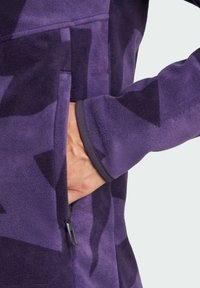Chaqueta de forro polar morada con un bolsillo con cremallera y detalles grises en los puños. El material tiene una textura suave y un patrón geométrico.
