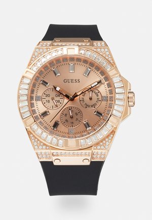 Montre en or rose avec un bracelet en caoutchouc noir, des accents en cristal et un cadran brun comportant trois sous-cadrans et des index horaires rectangulaires.