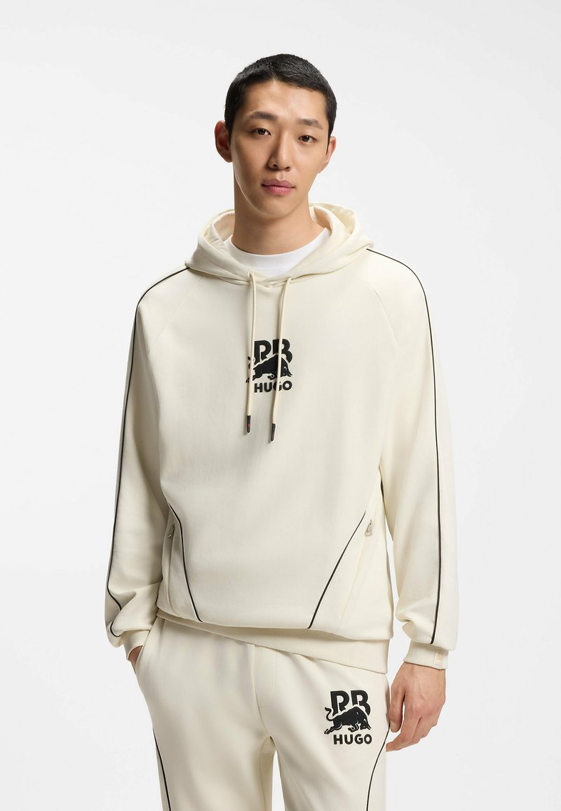 Jeune homme portant un sweat à capuche crème et un pantalon de survêtement avec un liseré noir et un logo RB HUGO noir sur la poitrine et la cuisse.
