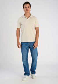 Ljusbeige kortärmad pikétröja med ett strukturerat mönster, kombinerad med blå denimjeans och vita sneakers. En enkel bakgrund.