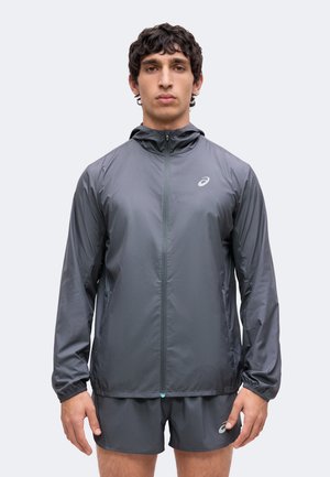 Homme portant une veste à capuche grise zippée et un short assorti, debout devant un fond clair uni.