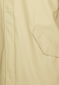 Beige regnrock med en mjuk textur, med en diagonal framfickficka och subtila sömnadsdetaljer på frampanelen.