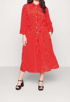 Femme portant une robe midi rouge à rayures verticales blanches, boutonnée sur le devant, taille avec cordon, manches 3/4, et talons blocs noirs avec bride à la cheville.