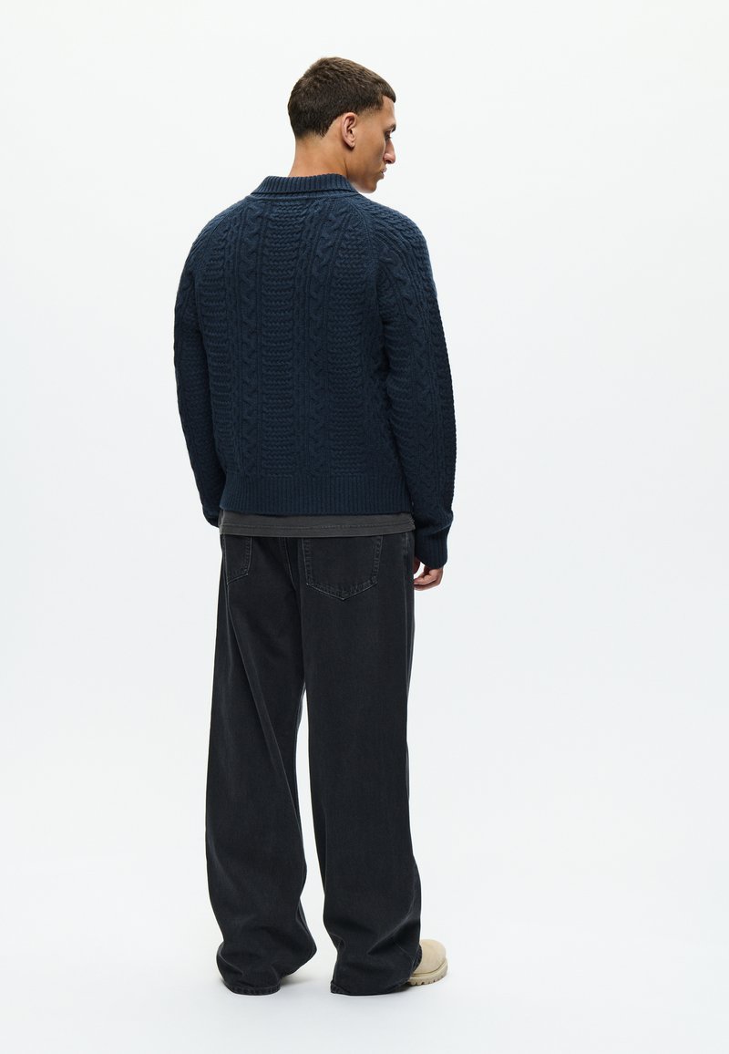 Marineblauer Kabelstrickpullover mit hohem Kragen, gerippten Bündchen und Saum. Getragen über einem grauen Hemd. Kombiniert mit schwarzen weit geschnittenen Jeans und beigen Schuhen.
