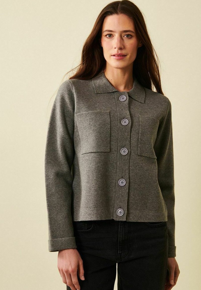 Nainen, jolla on harmaa napillinen neuletakki, jossa on kaksi etutaskua, ja mustat housut, yksiväristä beigeä taustaa vasten.