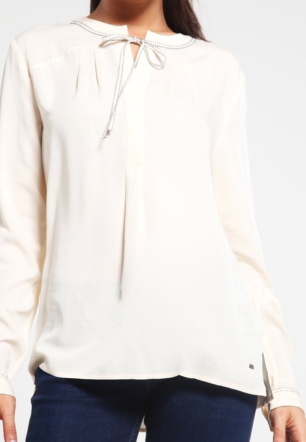 Blouse blanche ample à manches longues avec un détail noué à l'avant, portée avec un jean bleu foncé sur une personne sur un fond uni.