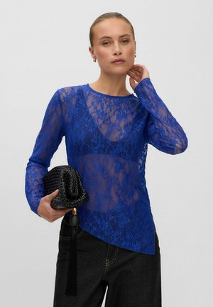 Femme portant un haut en dentelle florale bleue transparente par-dessus un bralette assorti, tenant une pochette noire tissée avec un pompon, posant devant un fond uni.
