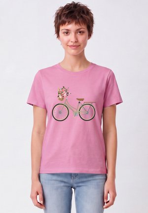 Jeune femme aux cheveux courts portant un t-shirt rose avec un motif de vélo vert et un panier de fleurs, assorti à un jean bleu clair.