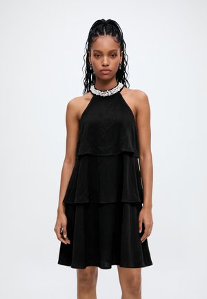 Femme aux cheveux tressés portant une robe noire sans manches à volants avec un col en perles sur un fond clair uni.