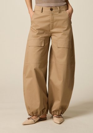 Pantaloni cargo beige a gamba larga con tasche a patta e polsini elasticizzati, indossati con ballerine beige con punta a punta e cinturini decorativi.