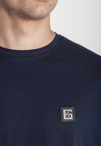 Detailný záber na osobu so tmavomodrým tričkom, na ktorom je malá štvorcová nášivka s prepojenými písmenami "K" a "G" na hrudi.