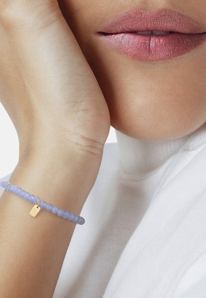 Primer plano de la parte inferior del rostro de una persona con labios naturales, apoyando la mano en la mejilla, llevando una pulsera de cuentas morado claro con un charm dorado.