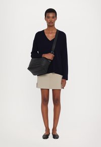 Geantă de umăr din piele neagră cu un finisaj texturat, purtată în stil crossbody. Model îmbrăcat cu un pulover bleu cu decolteu în V, fustă bej și balerini negri.