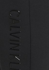 Svart sportleggings med en glänsande svart "CALVIN KLEIN"-logga som löper vertikalt längs sidan, tillverkade av mjukt, stretcht tyg.