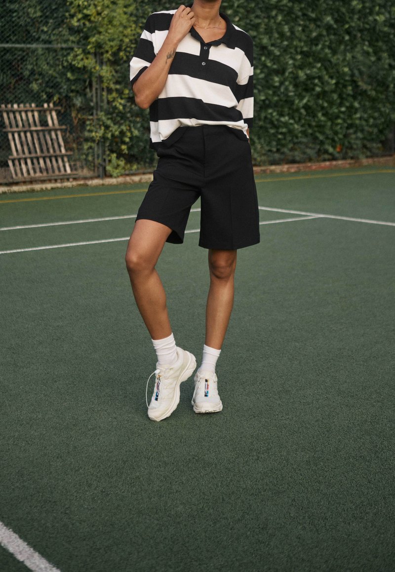 Shorts neri abbinati a una polo a righe bianche e nere. Sneakers bianche con accenti colorati e calzini bianchi su un campo da tennis verde.