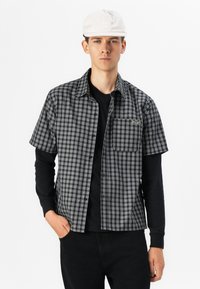 Short-sleeve plaid shirt in black and white with a chest pocket. Worn over a black long-sleeve shirt, paired with black pants and a white cap.