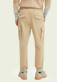 Bagudsig af en person iført beige cargo bukser med klaplommer og lysegrå sneakers med orange såler.