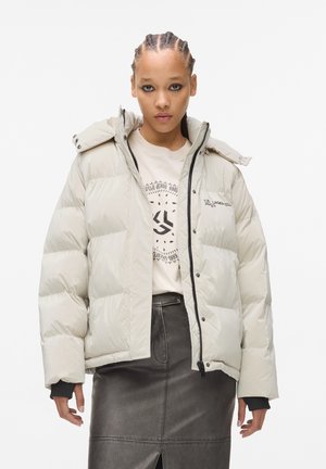 Dikke witte jas met een hoge kraag, rits aan de voorkant en drukknoopjes. Gekleed over een beige graphic t-shirt en een zwarte leren rok.