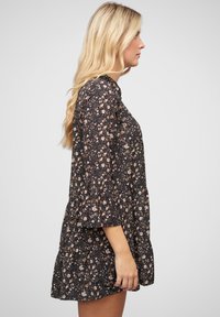 Robe noire à fleurs avec des manches longues et évasées et un design en étages. Présente de petites fleurs pêche et crème, fabriquée en tissu léger.