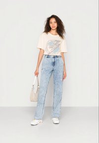 T-shirt gráfica bege com logótipo, jeans claros de cintura alta, ténis brancos e uma pequena mala bege, em pé contra um fundo neutro.