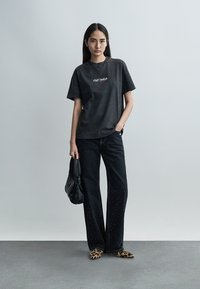 Μια γυναίκα στέκεται φορώντας μια ξεθωριασμένη μαύρη μπλούζα Off-White, φαρδιά μαύρα τζιν με στρας, μπαλαρίνες με λεοπάρ σχέδιο, και κρατώντας μια μαύρη τσάντα χειρός.