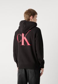 Melna kapuce ar rozā "CK" logo aizmugurē. Iespējota ar aukliņu regulējamu kapuci, ribotām aprocēm un mīkstu auduma struktūru. Valkāts ar tumšām džinsām.