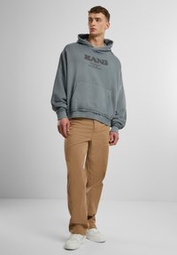 Gray oversized hoodie with a front pocket and printed logo, paired with tan straight-leg pants and white sneakers. Smooth fabric texture.