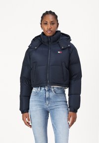Marineblå pufferjakke med kort design, med glidelås foran, trykknapper, hette og en liten logo-patch på brystet.