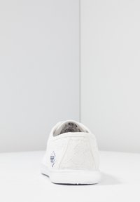 Sneaker blanc avec une empeigne en lacet texturée, semelle en caoutchouc et logo de marque noir sur le talon. Forme arrondie avec un design à tige basse.
