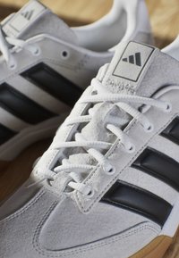 Weiße Wildleder-Sneaker mit schwarzen Streifen, weißen Schnürsenkeln und Gummisohlen auf einer Holzoberfläche.