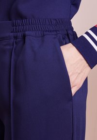 Navy urheiluhousut, joissa on teksturoitu pinta, vartalonmyötäinen vyötärö ja sivutasku. Hihassa on raidallinen yksityiskohta punaisena ja valkoisena.