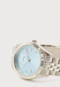 Sudraba metāla Timex rokas pulkstenis ar gaiši zilu ciparnīcu, sudraba stundu marķieriem un metāla saišu jostu, kurā laiks ir aptuveni 7:56.