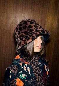 Pălărie de tip bucket cu imprimeu leopard maro, din blană artificială, având o margine lată și curbată. Interiorul are o căptușeală neagră. Stilizată cu o jachetă cu model.