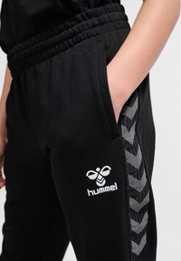 Schwarze Jogginghose mit elastischem Bund und Seitentaschen. Mit einem weißen Logo und grauen Chevron-Streifen an der Seite. Weicher Stoff.