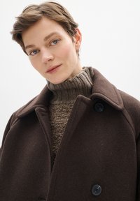 Abrigo de lana marrón con grandes solapas y botones negros, superpuesto a un suéter de punto texturizado marrón claro con cuello alto.