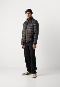 Sort pufferjakke med lodret quiltet design, parret med en olivengrøn top, sorte bukser og brune/grå sneakers med striber.