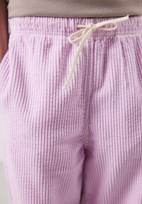 Nahaufnahme von lavendelfarbenen, gerippten Stoffhosen mit elastischem Bund und beigefarbenem Kordelzug, der zu einer Schleife gebunden ist.