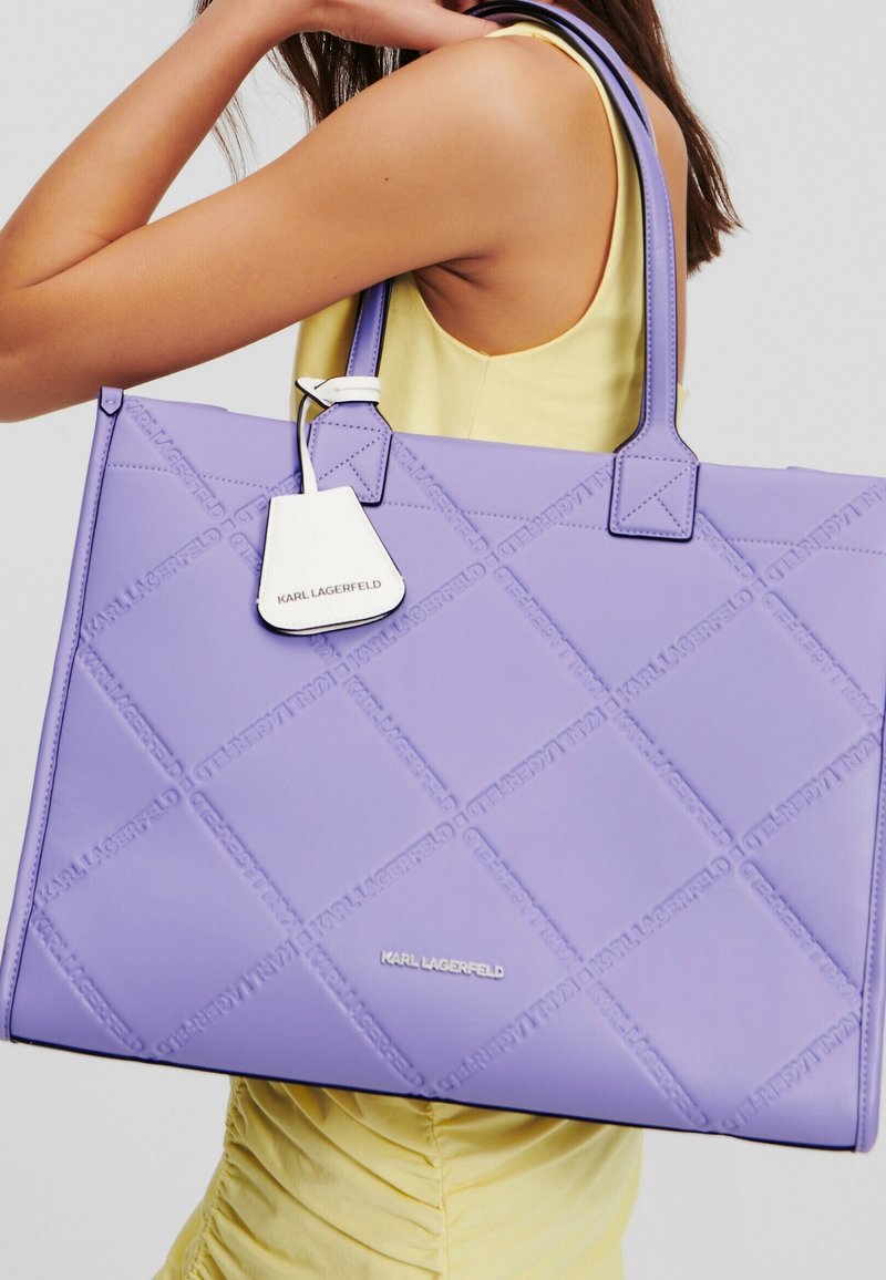 Sac fourre-tout matelassé lavande avec des coutures marquées, deux poignées et une étiquette blanche affichant "KARL LAGERFELD". Matière synthétique lisse.